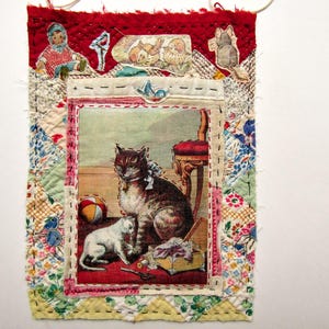 May include: A rectangular fabric wall hanging featuring a central printed image of a brown tabby cat and a white kitten. The border is composed of various quilted fabric patches in red, yellow, and floral patterns, with small cut-out figures stitched on top.