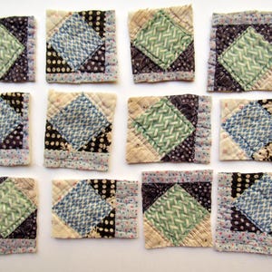 May include: Twelve square fabric quilt blocks arranged in a grid. Each block features a central diamond pattern in light green or blue, surrounded by various dark blue, black, and cream-colored patterned fabric borders with visible quilting stitches.