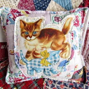 May include: A decorative pillow featuring a vintage-style illustration of a ginger kitten with blue eyes and a blue gingham kitten. The pillow is surrounded by a patchwork design with floral and patterned fabrics, creating a cozy, handcrafted aesthetic.
