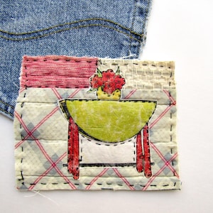 May include: A fabric patch featuring a green table with red legs and a bowl of red flowers. The patch is sewn onto a patterned fabric with red plaid lines and a denim background.