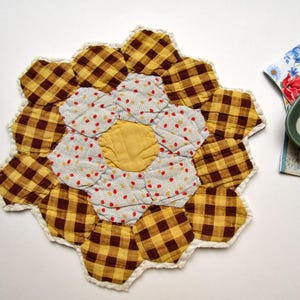 May include: A quilted fabric mat shaped like a flower, featuring a solid yellow center, a middle ring of light gray fabric with small red and yellow dots, and an outer border of yellow and dark brown checkered fabric with white trim edges.