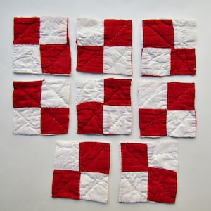 May include: Eight square fabric patches arranged in a grid, each featuring a four-patch checkerboard pattern in alternating red and white colors. The fabric has a quilted texture with visible diagonal stitching lines across the surface.
