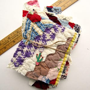 May include: A stack of handmade, patchwork coasters in various colors and patterns, tied with twine. The coasters feature a mix of fabrics, including floral, striped, and solid designs. A wooden ruler is placed diagonally across the coasters. A cactus charm is attached to the twine.