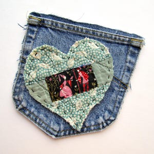 May include: A denim pocket with a heart-shaped patch. The patch is made of fabric with a floral pattern in shades of teal and green, and a central rectangle with figures in pink and red. The patch is stitched onto the pocket with black thread.