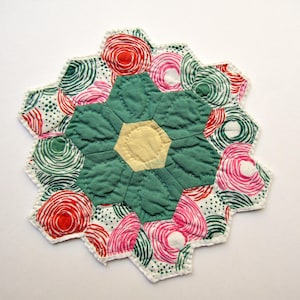 May include: A quilted fabric coaster featuring a central pale yellow hexagon surrounded by six dark teal petal-shaped pieces. The outer border consists of smaller hexagons with white backgrounds and circular patterns in red, pink, and dark green.