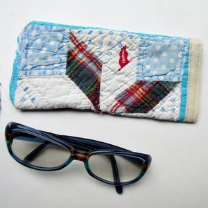 May include: A pair of blue eyeglasses with a patterned frame rests near a quilted glasses case. The case features a patchwork design with blue polka dots, plaid, and a red tag that reads "Tropi".