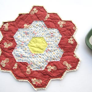 May include: A handmade, quilted hot pad in a flower shape. The center is yellow, surrounded by a ring of blue, pink, and white patterned fabric. The outer petals are red with a white floral pattern, and the edges are trimmed with a cream-colored ruffle.