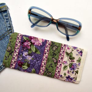 May include: A pair of blue and tortoiseshell eyeglasses positioned above a rectangular quilted fabric case featuring a floral pattern in shades of purple, green, and cream, alongside a corner of blue denim fabric.