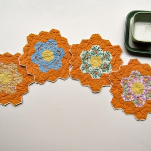 May include: Four quilted hexagonal coasters featuring a vibrant orange border and a central floral pattern in shades of yellow, blue, green, and purple. Each coaster has a textured, stitched surface and a white edge. A green square bowl sits nearby.