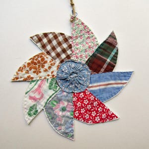 May include: A circular fabric wall hanging featuring eight wedge-shaped petals made from various patterned textiles, including brown gingham, floral prints, plaid, and stripes, surrounding a central blue gathered fabric circle with a twine loop.