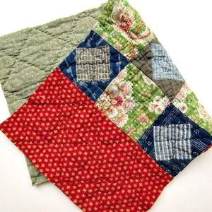 May include: A quilted fabric piece featuring a patchwork design with various patterns including red with gold stars, blue with white grid lines, and green with floral motifs. The reverse side is a light green fabric with small white dots.