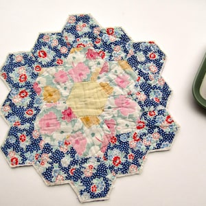 May include: A handmade, floral-patterned coaster with a hexagonal design. The coaster features a central yellow hexagon surrounded by pink, blue, and white floral print hexagons. The outer ring has a blue polka dot pattern with red and white flowers.