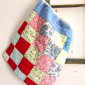 May include: A patchwork drawstring bag with a variety of fabric patterns and colors. The bag features red, light blue, floral, and paisley squares. The top is a solid blue fabric with a drawstring closure. The bag is hanging against a white wall.