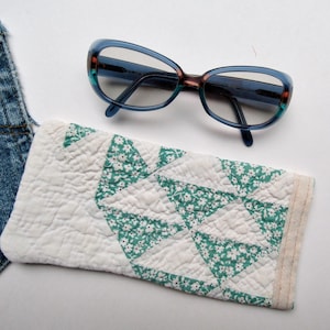 May include: A pair of blue-tinted glasses with a multi-colored frame resting above a rectangular quilted fabric case. The case features a white and teal floral pattern with a geometric stitched design and a light-colored fabric trim edge.