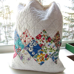 May include: A white quilted drawstring bag with a patchwork design. The patchwork features various floral patterns in red, blue, yellow, and green. The bag is made of quilted fabric and has a drawstring closure.