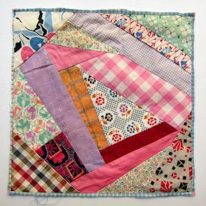 May include: A square patchwork quilt block featuring a variety of fabric patterns including pink gingham, floral prints, orange plaid, and geometric shapes. The fabric pieces are stitched together in a diagonal arrangement with a blue checkered border.