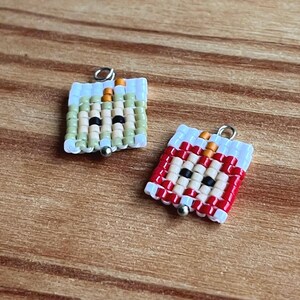 Op de afbeelding: Twee vierkante kralenhangers, één groen en beige, de andere rood en wit, elk met een zilverkleurige beugel en een kleine zilveren kraal. De hangers hebben een gezichtsontwerp met zwarte ogen en oranje accenten.