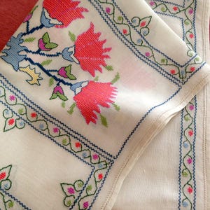 May include: A white fabric textile featuring intricate cross-stitch embroidery. The design includes large, vibrant red floral motifs with blue and green accents, complemented by a delicate green and blue vine border pattern along the edges.