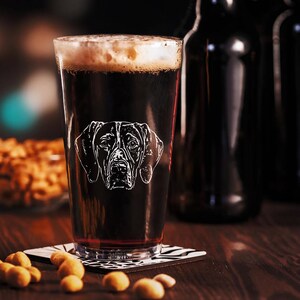May include: A pint glass filled with dark beer and foam, featuring a white etched design of a dog's face. The glass sits on a black and white patterned coaster, with peanuts scattered around it. Dark bottles are in the background.