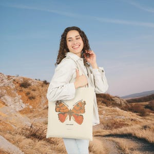 May include: Cream-colored tote bag featuring a detailed butterfly illustration in shades of orange and brown. The text "transformed in the renewing of your mind" is printed above and below the butterfly. The bag is held by a person.