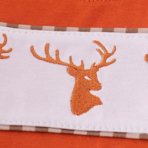 May include: Close-up of an orange shirt with a white and brown striped accent. The white accent features embroidered orange deer silhouettes. The shirt has a round neckline and is made of a soft material.