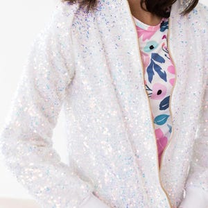 May include: A white sequined bomber jacket with a gold zipper. The jacket has white ribbed cuffs and a white hem. The interior lining of the jacket is visible, featuring a floral pattern with pink, blue, and white flowers.