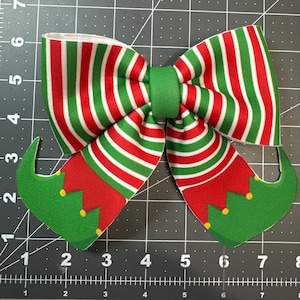May include: A red and green striped Christmas bow with elf boot accents. The bow is tied with a green ribbon and has a white background.