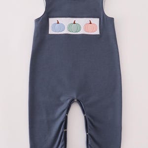 Puede incluir: Mono de bebé azul marino con ribete blanco y tres cierres de botón en cada hombro. El mono presenta un rectángulo bordado con tres calabazas en azul, verde y rosa.