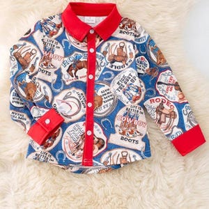 Pode incluir: Camisa abotoada com tema de cowboy. A camisa apresenta um fundo azul com gráficos e texto relacionados a cowboys, acentuada por uma gola e punhos vermelhos. Possui mangas compridas e fecho de botões.