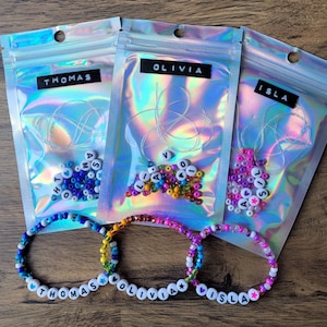 May include: Three personalised beaded bracelets with the names THOMAS, OLIVIA, and ISLA. Each bracelet features white letter beads and colourful accent beads. Three iridescent pouches contain beads and string, labelled with the same names.