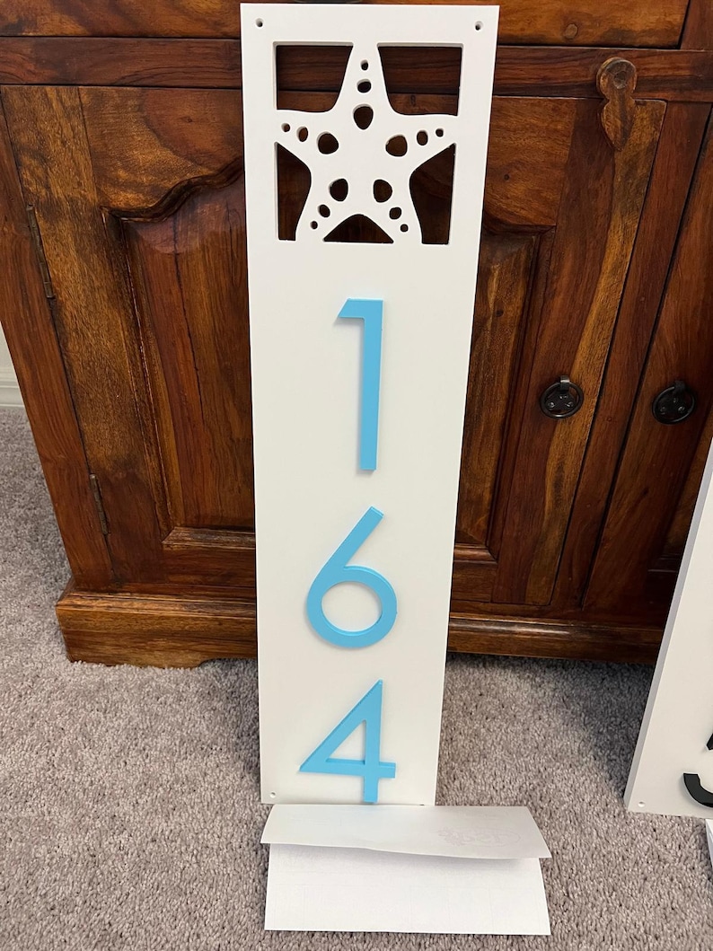 Puede incluir: Un letrero de madera blanco con un recorte de una estrella de mar y los n&uacute;meros 164 pintados en azul.