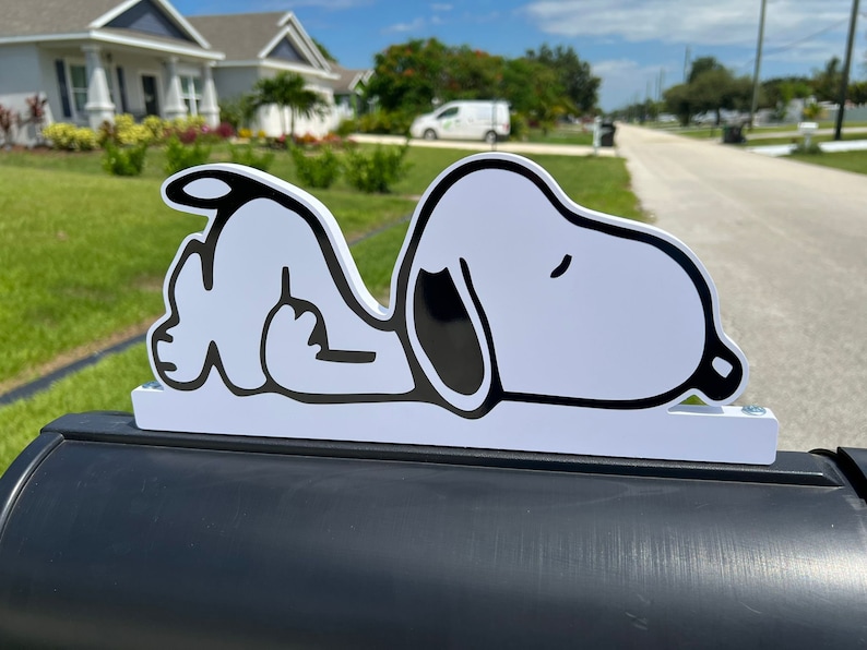 以下が含まれることがあります： 白地に黒と白のSnoopyのイラストが描かれた郵便受けの飾り。耐久性のあるプラスチック製で、ほとんどの標準的な郵便受けに合うように設計されています。
