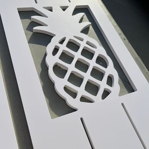 May include: White wooden sign with a cutout pineapple design. The pineapple is in the center of the sign and is surrounded by a white border.