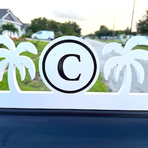 May include: White wooden mailbox topper with two palm tree silhouettes and a central circular design. The circle features a black letter "C" on a white background, framed by a black border. The topper is mounted on a dark surface.