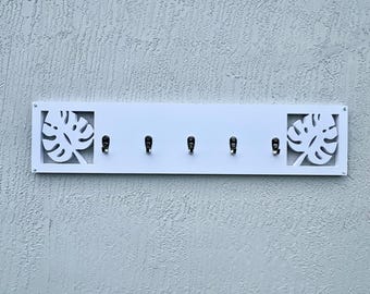 May include: White key holder with five black hooks. The key holder has a rectangular shape with decorative cut-out monstera leaves on each end. The key holder is mounted on a textured, light gray wall.