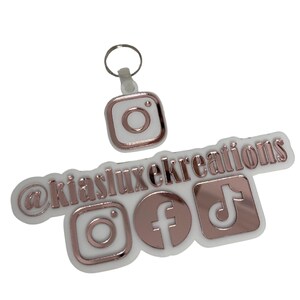May include: A white keychain featuring a rose gold Instagram logo and the text "@kialuxekreations". Below are rose gold logos for Instagram, Facebook, and TikTok. The keychain is made of a glossy material.