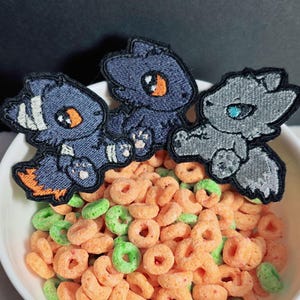 May include: Three embroidered patches featuring stylised cartoon creatures. Two patches are dark blue with orange eyes and white accents, while the third is light grey with a bright blue eye. Each patch has a black border and sits in a white bowl.