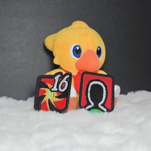 May include: A yellow plush bird toy with a large orange beak and blue eyes, positioned behind two square embroidered patches. The left patch features a red and yellow starburst design with the number 16, and the right patch shows a black silhouette.