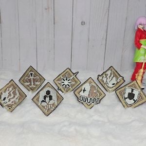 以下が含まれることがあります： さまざまなシンボルとデザインが施された、7つの刺繍パッチのコレクション。 各ダイヤモンド型のパッチは、金色の縁とベージュの背景を持っています。 パッチには、天秤、スターバースト、その他の抽象的なデザインの画像が表示されています。