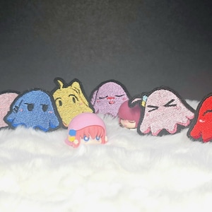 以下が含まれることがあります： ピンク、青、黄色、赤の様々なゴーストのようなキャラクターが刺繍されたワッペンと、ピンクの髪と帽子を身につけた小さなフィギュアが、白い質感のある表面にグループとして配置されています。