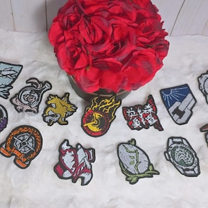 Puede incluir: Una colección de parches bordados con varios diseños, incluyendo una brújula, un sol sobre una montaña y texto japonés. Los parches presentan una variedad de colores, incluyendo azul, naranja, amarillo y rojo, y están dispuestos sobre una superficie blanca texturizada.