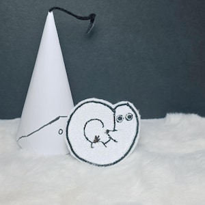Könnte beinhalten: Ein weißer, kegelförmiger Partyhut mit einer schwarzen Strichzeichnung und einem schwarzen flauschigen Aufsatz, neben einem runden, weißen Stickaufnäher mit einer schwarzen Strichzeichnung eines kleinen Wesens mit großen Augen und geringeltem Schwanz.