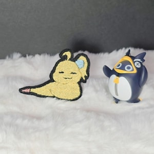 Könnte beinhalten: Ein gelber gestickter Aufnäher mit einer stilisierten Kreatur mit schwarzem Rand und einem kleinen blauen Akzent, platziert neben einer dunkelblau-weißen pinguinartigen Figur mit gelbem Schnabel und einem Reißverschlussdetail.
