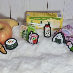 May include: An assortment of embroidered food patches, including a carrot, sushi roll, strawberry drink, and onigiri. A yellow box of juice, a red apple, and a banana are also present. The patches are on a white, fluffy surface.