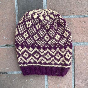 May include: A knitted beanie hat in a diamond pattern. The hat features a deep purple brim and a cream and purple geometric design across the crown. The hat is made of a soft, warm material, perfect for cold weather.