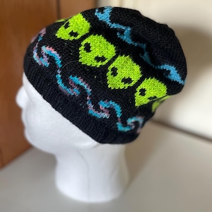 May include: Black knit beanie featuring neon green alien faces and blue and pink wavy designs. The beanie is displayed on a white mannequin head. The hat is a winter accessory.