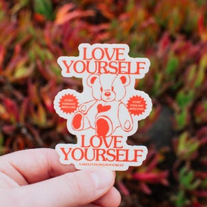 Może przedstawiać: Biała naklejka z czerwonym obramowaniem przedstawia kreskówkowego misia trzymającego czerwone serce. Naklejka głosi "LOVE YOURSELF" dużymi czerwonymi literami i "START YOUR DAY WITH LOVE" mniejszymi czerwonymi literami. Naklejka zawiera również tekst "@SELFLOVINGMOVEMENT".