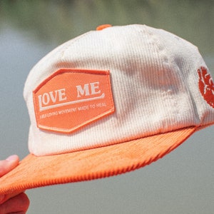 May include: A white corduroy baseball cap with an orange brim. The front of the cap has a patch that says "LOVE ME A HEALING MOVEMENT MADE TO HEAL". The back of the cap has a small orange heart.