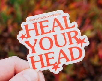 May include: White sticker with the text "HEAL YOUR HEAD" in orange letters with stars on either side. The sticker is held in a hand against a blurred background of red and green foliage. The text "@ASELFLOVINGMOVEMENT" is in smaller letters at the top of the sticker.