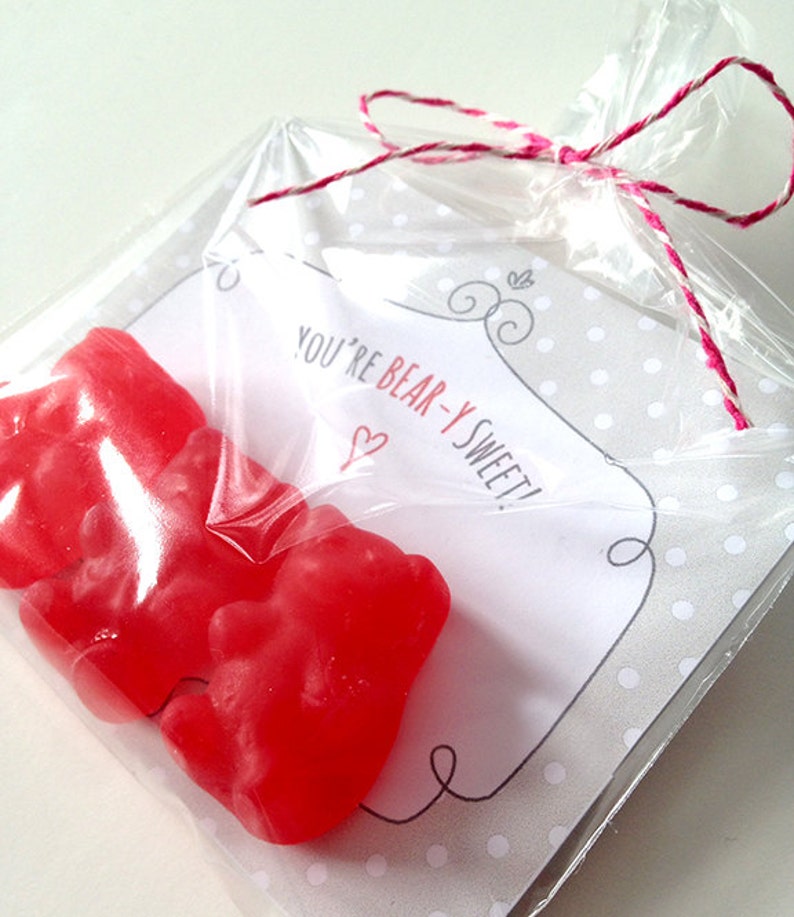 May include: A clear plastic bag tied with pink and white twine contains two red gummy bears and a white card with the text "You're Bear-y Sweet!" printed in black with a small red heart.